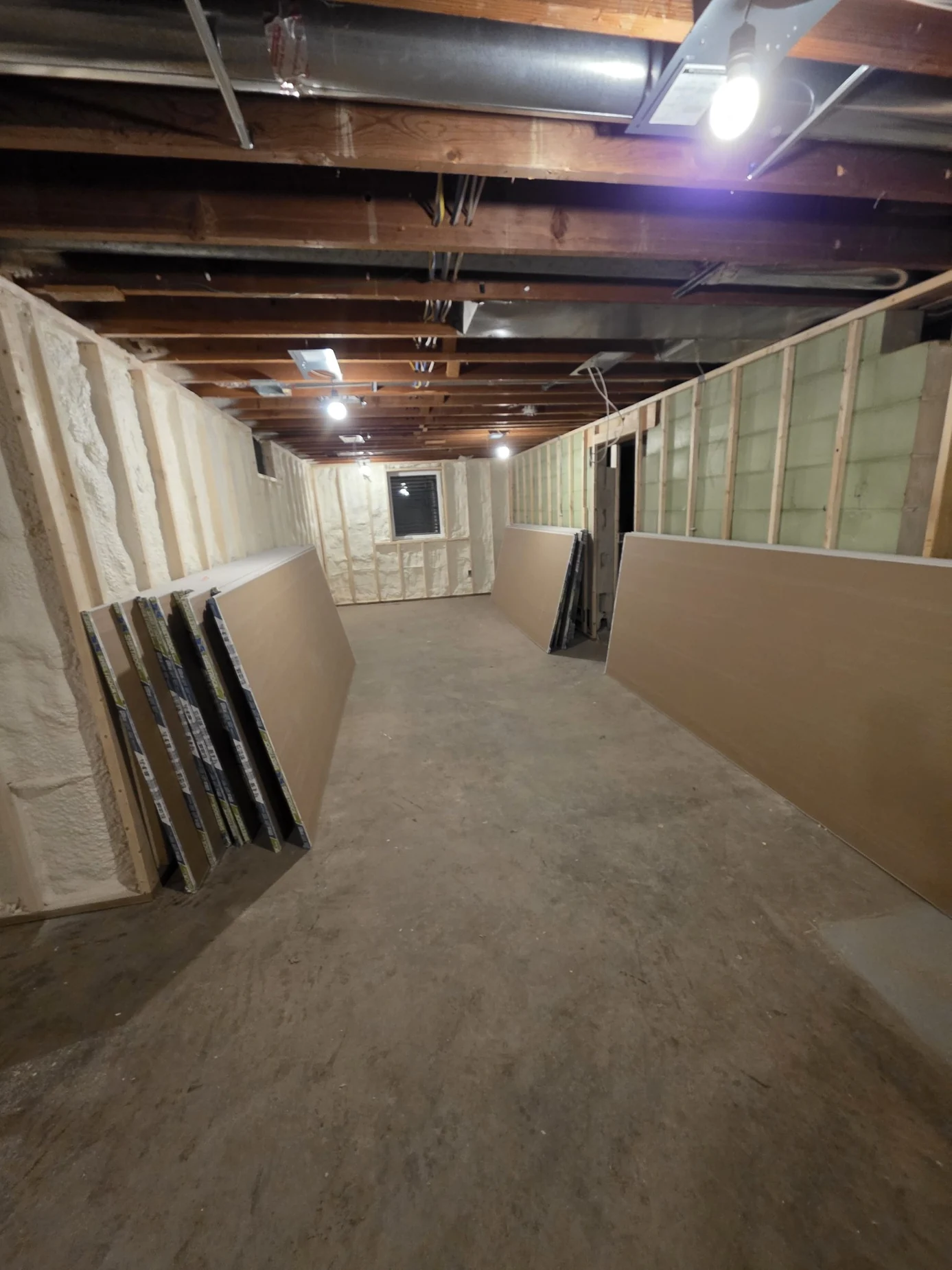 Same basement room with insulated framing and drywall panels stacked along the walls