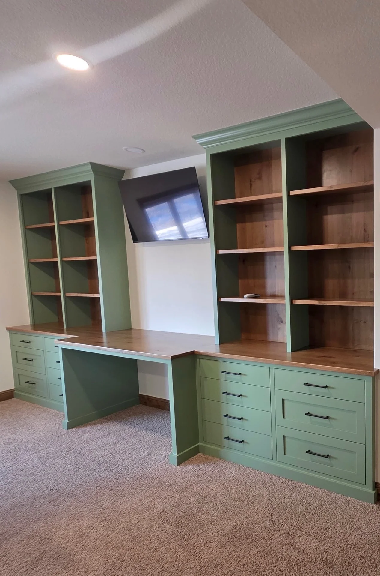 Completed sage-green built-in shelving and desk with wood countertops and black drawer handles