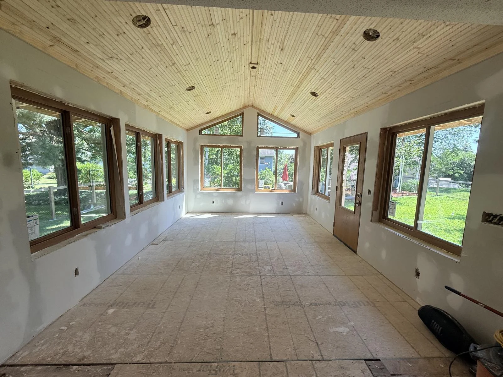 Sunroom renovation with bare subfloor and unfinished newly boarded walls below a wood ceiling